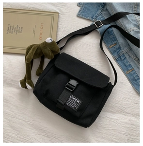 Detail of black canvas messenger bag featuring plastic buckle and a green frog plush charm