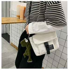 Detail of white canvas messenger bag with black buckle and plush frog charm accessory