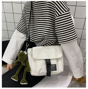 Close-up of white canvas messenger bag with black buckle and green frog plush charm
