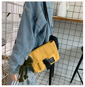 Detail view of yellow canvas messenger bag featuring a green frog plush charm and buckle