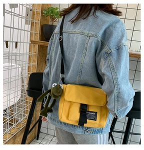 Detail of yellow canvas messenger bag with black buckle and plush frog pendant accessory