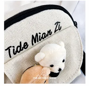 Detail of off-white canvas crossbody bag with black embroidery and cute plush bear charm