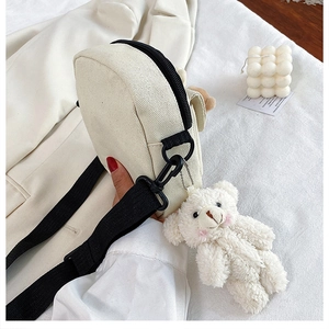 Detail of off-white canvas crossbody bag with black strap and a cute plush bear pendant
