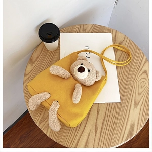 Close-up of yellow canvas shoulder bag with a plush 3D teddy bear charm on wood surface