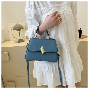 Close-up of a blue trapezoidal handbag with a golden S-clasp held by a model in a dress
