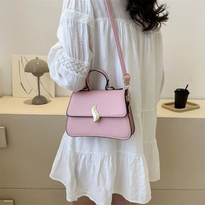 Pink trapezoid crossbody bag with gold buckle worn by a model in a minimalist room
