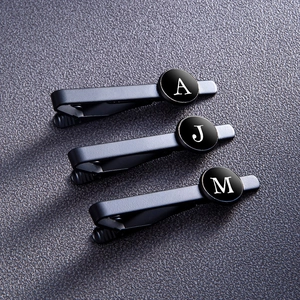 Three matte black metal tie clips with white initials A, J, and M on a grey leather background