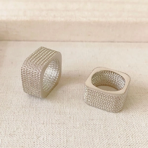 Minimalist square silver rings with textured woven pattern on a beige fabric background