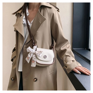 Detail of crossbody saddle bag with metal logo and floral ribbon on beige trench coat