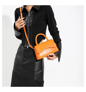 Model holding a vibrant orange leather handbag with wavy flap and adjustable shoulder strap
