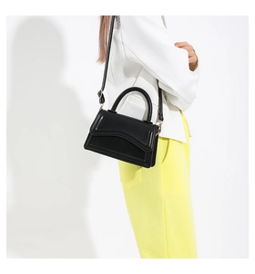 Detail of a black crossbody bag featuring a unique wavy flap and top handle on a model