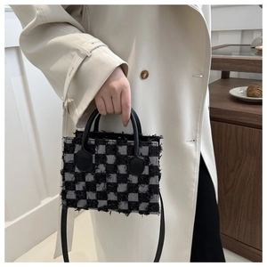 Detail of checkered denim handbag with fringe edges and leather handles on beige coat