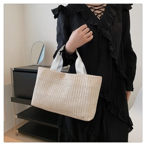 Woman holding a beige woven straw tote bag with white ribbon handles in a modern room