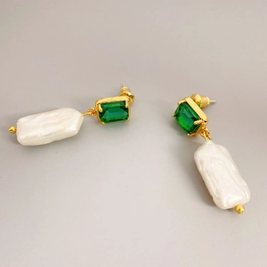 Macro shot of long baroque pearl earrings with emerald-cut green gemstones on gray surface