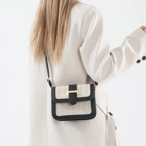 Woven straw crossbody bag with black leather trim and gold buckle on a white blazer