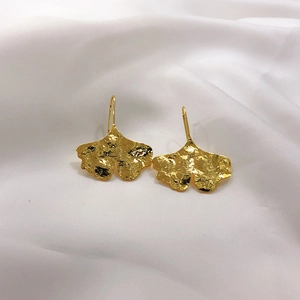 Macro shot of gold-plated ginkgo leaf earrings with hammered texture on white silk fabric