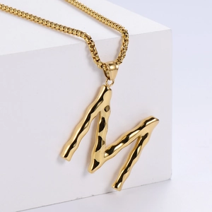 Détail du pendentif lettre M doré avec texture ondulée sur un support blanc pur