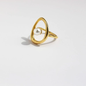 Bague artistique dorée avec perle blanche suspendue sur un fond gris clair minimal