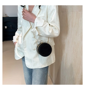 Detail of a black velvet round box bag with gold chain strap on a white textured blazer