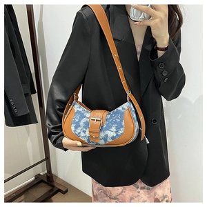 Detail of blue denim saddle bag with brown leather trim and large metal buckle shown on model