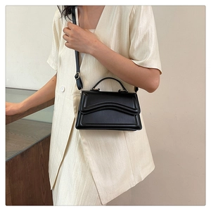 Detail of a chic black trapezoid handbag with wavy flap design on a model