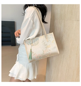 On-shoulder view of an off-white silk tote bag with floral patterns and a green tassel