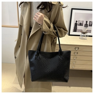 Detail of a black woven faux leather tote bag carried by a model in a beige trench coat
