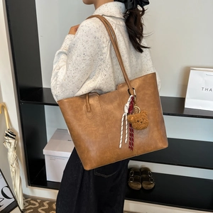 Brown faux leather tote bag with teddy bear charm and woven rope on simple display