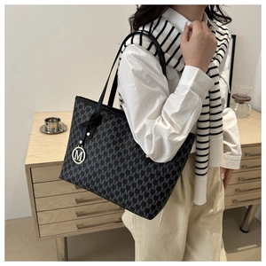 Detail of black tote bag with gray M-pattern, gold charm, and slim straps on model