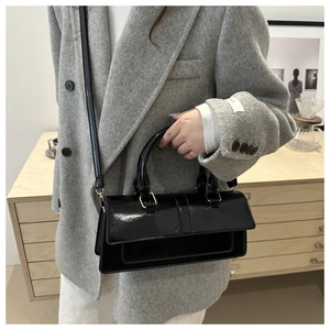 Glossy black patent leather baguette bag with gold buckle shown on shoulder of a model