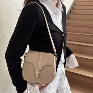 Minimalist beige saddle shoulder bag with gold clasp shown on a model in a cozy indoor setting