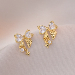 Golden bow stud earrings with heart zirconia and star charms on soft fabric background