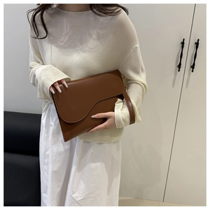 Brown leather envelope bag with asymmetrical curved flap held by hand in a minimalist setting