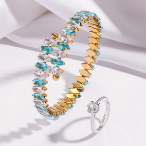 Close-up of gold bangle with blue marquise zircon and a crystal ring on white silk