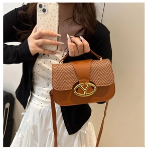 Detail of a brown leather saddle bag featuring a woven flap and gold oval logo buckle