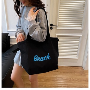 Detail of a large black canvas tote bag with blue Beach embroidery worn by a model