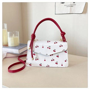 Detail of white leather shoulder bag with red cherry prints and red handle on a desk