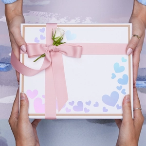 Close-up of decorative gift box with pink satin ribbon and white rose held by hands