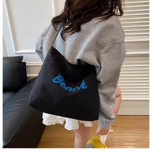 Προβολή lifestyle μιας μεγάλης μαύρης τσάντας tote με μπλε γράμματα Beach στον ώμο