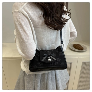 Schwarze Schultertasche mit Schleife und Herzanhänger an Model vor schlichtem Hintergrund