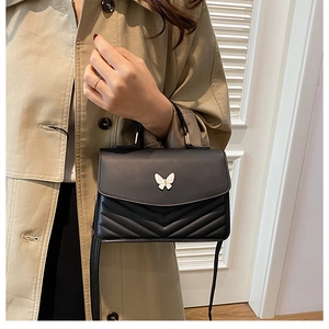 Detail of a black quilted crossbody bag with butterfly clasp on a beige trench coat