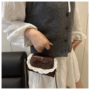 Detail of a dark brown patent leather mini bag with lace trim and bow on a model