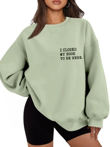 Close-up of sage green sweatshirt featuring 'I CLOSED MY BOOK TO BE HERE' printed text