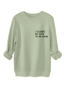 Close-up of ribbed crewneck and 'I CLOSED MY BOOK TO BE HERE' print on green sweatshirt