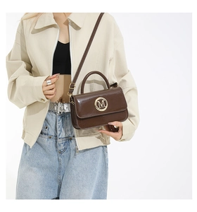 Dettaglio borsa a tracolla in vernice marrone scuro con logo M dorato su outfit beige