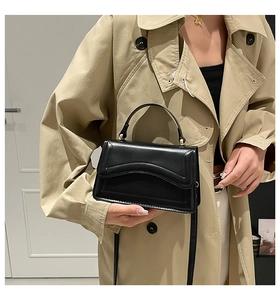 Detail of a chic black glossy leather crossbody bag with curved stitching on a trench coat