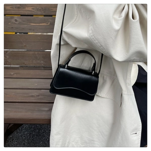 Model wearing a black wavy-flap crossbody bag over a white coat on a wooden bench