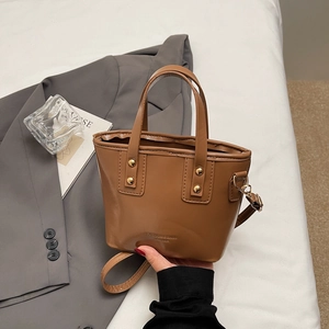 Brown leather trapezoid crossbody bag with top handles and golden studs on gray suit background