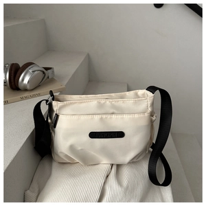 Minimalist beige nylon crossbody bag with wide black strap next to fashion magazine