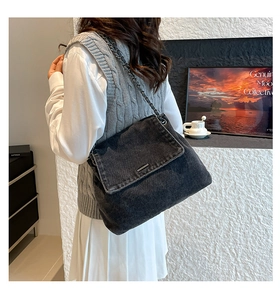 Detail of a black denim shoulder bag with metal chain strap worn by a model in a room